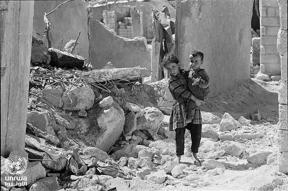 A girl carries a toddler through the ruins of a refugee camp